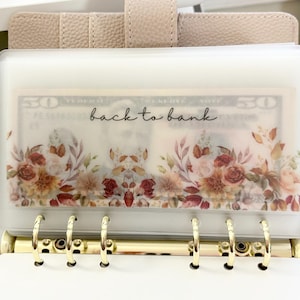 May include: A beige binder with gold-colored rings and a clear pocket. The pocket displays a floral design with the text "back to bank" in black script. The design includes roses and leaves in shades of red, brown, and beige.