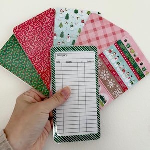 May include: A hand holds a set of Christmas-themed budget envelopes and a matching budget sheet. The envelopes feature patterns of candy canes, snowmen, and plaid. The budget sheet has a green border and space for category, date, details, and balance.