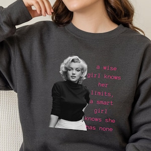 May include: Dark heather gray sweatshirt featuring a black and white image of Marilyn Monroe. The sweatshirt has the text "a wise girl knows her limits, a smart girl knows she has none" in pink.