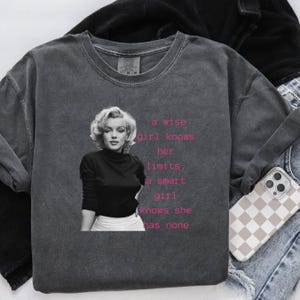 May include: Dark gray long-sleeve sweatshirt featuring a black and white image of Marilyn Monroe. The sweatshirt has the text "a wise girl knows her limits, a smart girl knows she has none" in pink.