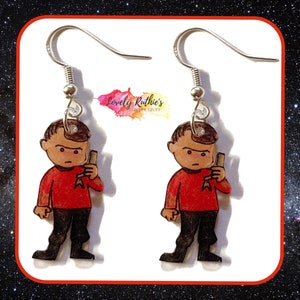 Puede incluir: Un par de pendientes con un personaje de dibujos animados con una camisa roja, pantalones negros y un phaser. El personaje es de la franquicia Star Trek.