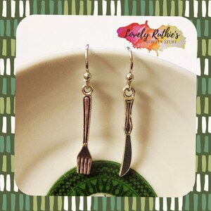 May include: Silver fork and knife earrings on a white plate with a green and white patterned design. The earrings are hanging from silver hooks. The plate is on a white surface. The text "Lovely Ruthie's Lovely Stuff" is visible in the top right corner of the image.