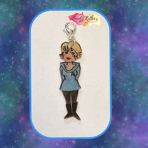 Puede incluir: Un charm plateado con una imagen de dibujos animados de una mujer con el pelo corto rubio que lleva un uniforme azul. El charm est&aacute; unido a un cierre plateado.