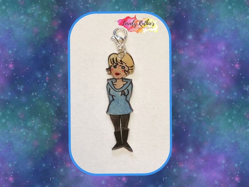 Puede incluir: Un charm plateado con una imagen de dibujos animados de una mujer con el pelo corto rubio que lleva un uniforme azul. El charm est&aacute; unido a un cierre plateado.