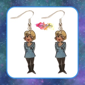 Puede incluir: Un par de pendientes con una ilustraci&oacute;n de dibujos animados de una mujer con un uniforme azul. Los pendientes son plateados y tienen un cierre de gancho.