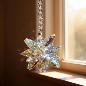 May include: A clear, faceted crystal suncatcher with a starburst design. The suncatcher is suspended by a string of clear beads. The crystal reflects light, creating a rainbow effect. The suncatcher is hanging near a window.