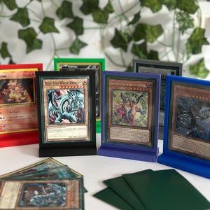 Op de afbeelding: Een display van verzamelkaarten, waaronder een "Blue-Eyes White Dragon" kaart, in verschillende gekleurde stands. Groene kaarthoezen zijn zichtbaar op de voorgrond. De achtergrond toont kunstmatig klimop.