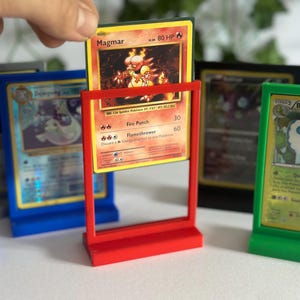 Op de afbeelding: Een rode displaystandaard met een Magmar Pokémon-kaart. De kaart heeft een gele rand en bevat een illustratie van een vurige creatuur. Andere Pokémon-kaarten zijn zichtbaar in blauwe, groene en zwarte standaards.