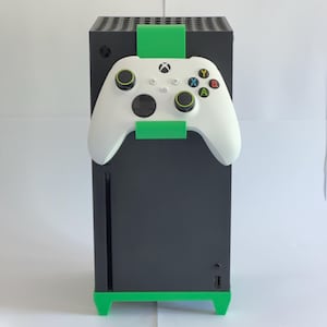 May include: A black games console with a white controller attached. The controller has coloured buttons and a green bracket holding it in place. The console has a green base and a green bracket at the top.