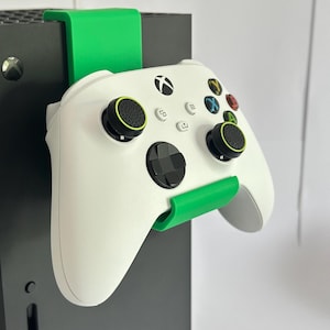 Może przedstawiać: Biały kontroler Xbox z czarnymi i zielonymi akcentami jest trzymany przez zielony wspornik przymocowany do czarnej konsoli. Kontroler ma kolorowe przyciski i czarne joysticki z zielonymi obwódkami.