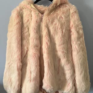 May include: A light pink faux fur hooded jacket with a zipper closure. The jacket is hanging on a black hanger against a light blue wall. The jacket has a drawstring at the bottom and a soft, fluffy texture.