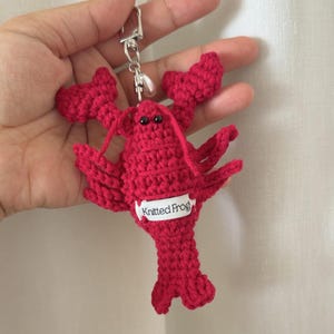 Puede incluir: Un llavero de langosta roja tejido a ganchillo. La langosta tiene ojos negros y una pequeña etiqueta blanca que dice "Knitted Frog". El llavero está sujeto a un cierre metálico plateado.