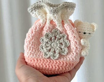 May include: A handmade crochet drawstring bag in shades of pink, white, and gray, featuring a gray flower applique and a small white teddy bear. The bag is held in a hand, showcasing its soft texture and detailed craftsmanship.
