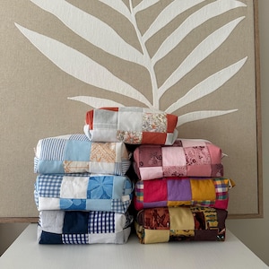 Könnte beinhalten: Ein Stapel bunter Patchwork-Stoffbeutel. Jeder Beutel ist rechteckig und weist eine einzigartige Kombination aus gemusterten Stoffen in verschiedenen Farben auf, darunter Blau, Rosa und Gelb. Die Beutel sind auf einer weißen Oberfläche gestapelt.
