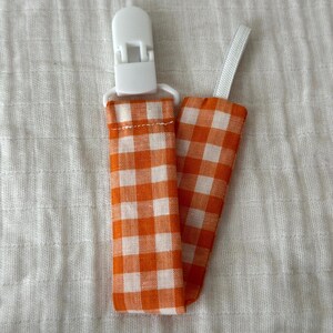 May include: A dummy clip featuring a fabric strap with an orange and white gingham check pattern. The strap is attached to a white plastic clip at one end and includes a small white elastic loop at the other end for secure attachment.
