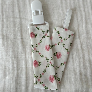 May include: A fabric dummy clip featuring a cream-coloured background with a repeating pattern of pink roses and green vines arranged in a diamond grid. The accessory includes a white plastic clip at the top and a white elastic loop at the end.