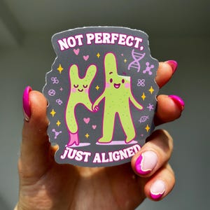 May include: A sticker featuring the text "NOT PERFECT, JUST ALIGNED" and two green cartoon figures holding hands. The sticker has a grey background with pink hearts and yellow stars.