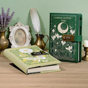 May include: Two green journals with combination locks. One upright journal features a crescent moon, floral motifs, and the text CANDLE-LIGHTIN' TIME. The other lying journal displays a large white daisy and butterfly illustration with intricate green foliage.
