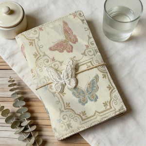 May include: A rectangular notebook cover featuring a cream-coloured fabric with a woven pattern of pink and blue butterflies and gold scrollwork. A white embroidered butterfly applique is attached to the centre by a thin elastic band.