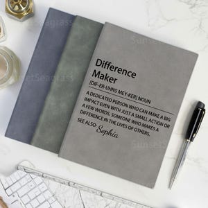 Könnte beinhalten: Drei Notizbücher in Grau- und Blautönen werden gezeigt, mit einem schwarzen Stift. Das vordere Notizbuch trägt den Text "Difference Maker" mit einer Definition und dem Namen "Sophia".