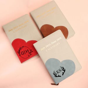 Puede incluir: Varios calendarios personalizados con detalles en forma de corazón. Los calendarios son de color beige claro con letras doradas que dicen "July 2025-June 2026 CALENDARS". Uno tiene un corazón rojo, otro un corazón marrón y el último un corazón azul.