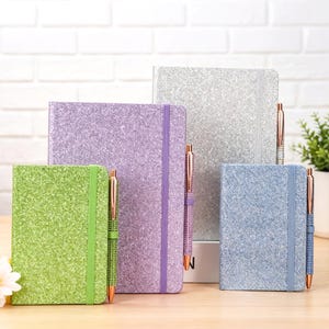 May include: A collection of glitter-covered notebooks in green, purple, silver, and blue, each with a matching elastic band and a rose gold pen. The notebooks are arranged on a wooden surface, with a white brick wall in the background.