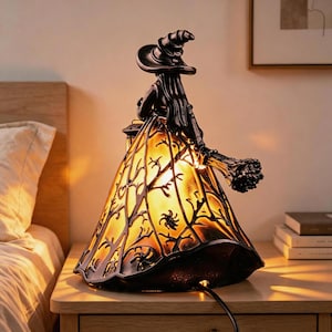 Könnte beinhalten: Eine schwarze Hexen-Figurenlampe mit spitzem Hut und langem Haar, die ein weites, ausgestelltes Kleid trägt, das in warmem Gelb leuchtet. Das Kleid zeigt ein dunkles, kompliziertes Überlagerungsmuster aus kahlen Ästen und Spinnennetz-Designs.
