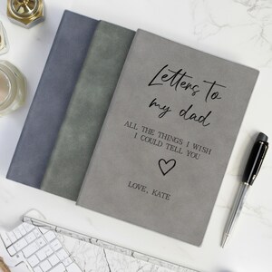 Pode incluir: Três cadernos em tons de azul e cinza. O caderno da frente é cinza e tem o texto "Letters to my dad" com um coração e as palavras "Love, Kate". Uma caneta preta e prateada está à direita.