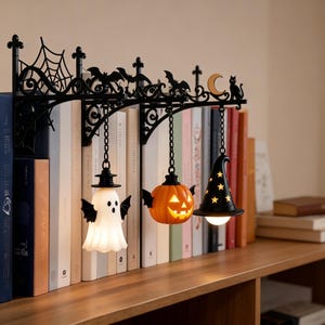 May include: A black metal shelf bracket featuring decorative cutouts of a spider web, bats, a crescent moon, and a cat. Three hanging ornaments are suspended from the bracket: a white ghost with wings, an orange jack-o'-lantern, and a black witch hat.