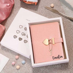 May include: A pink journal with a heart-shaped clasp and a small key charm, presented in a white box. The journal is personalized with the name "Emilia" in black script. The box is decorated with heart-shaped designs.