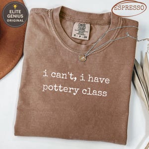 May include: Espresso-colored Comfort Colors t-shirt with the text "i can't, i have pottery class" in white. A silver necklace with a small pendant rests on the shirt. The shirt is folded on a white surface.