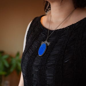 Könnte beinhalten: Eine silberne Halskette mit Anhänger, die einen großen, ovalen, leuchtend blauen Stein zeigt. Der Stein ist in einen silbernen Rahmen mit einem dekorativen kronenartigen Design gefasst. Die Halskette wird über einem schwarzen, strukturierten ärmellosen Oberteil getragen.