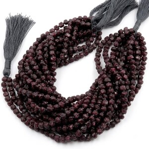 Peut inclure: Un long collier composé de perles bordeaux foncé, facettées et en forme de cube, disposées en plusieurs boucles. Le collier est terminé aux deux extrémités par des pompons en fil gris foncé, formant une boucle texturée de perles géométriques.
