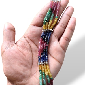 May include: A multi-strand gemstone bracelet with colourful beads. The bracelet includes strands of red, green, yellow, and blue gemstones, connected by silver clasps. The bracelet is displayed on a hand against a white background.