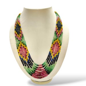 Peut inclure: Un collier de perles multicolores présenté sur un buste blanc. Le collier est composé de perles vertes, roses, jaunes, bleues et noires. Les perles sont disposées selon un motif géométrique.