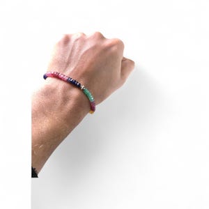 Könnte beinhalten: Ein mehrfarbiges Perlenarmband mit roten, rosa, blauen, grünen und gelben Edelsteinen. Das Armband ist auf einer dünnen Schnur aufgefädelt und hat einen kleinen silbernen Akzent. Das Armband wird an einem Handgelenk vor weißem Hintergrund getragen.