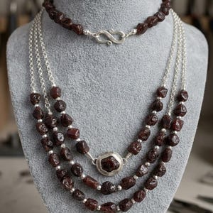 May include: A multi-strand necklace featuring dark red, irregular, tumbled gemstone beads interspersed with small, round, metallic silver-toned beads. The design includes a central circular silver-toned pendant and a silver-toned S-hook clasp.