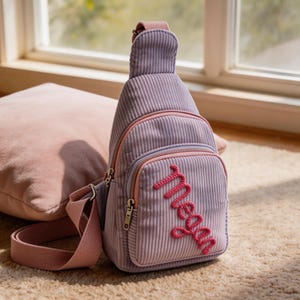 May include: A lilac corduroy sling bag with a pink embroidered name. The bag has a front zippered pocket, a top handle, and a pink adjustable strap. The bag is sitting on a beige carpet.