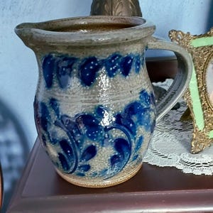 May include: A stoneware pitcher with a handle, featuring a blue floral design on a gray background. The pitcher has a wide opening and a spout for pouring. The handmade pottery is a decorative item.