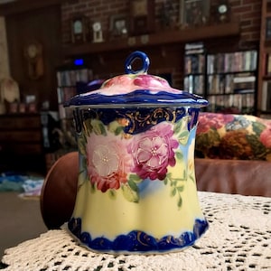 May include: A vintage porcelain jar with a blue and yellow floral design. The jar has a lid with a floral design and a gold trim. The jar is sitting on a white crocheted doily.