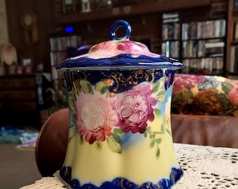 May include: A vintage porcelain jar with a blue and yellow floral design. The jar has a lid with a floral design and a gold trim. The jar is sitting on a white crocheted doily.