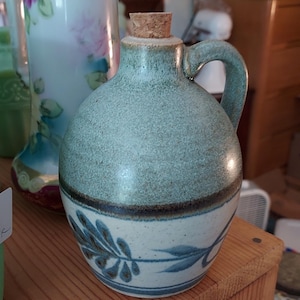May include: A small, round, ceramic jug with a handle and a cork stopper. The jug is primarily light blue with a textured surface and a white base decorated with blue leaf designs. The jug measures approximately 6 inches in height.