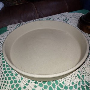 May include: A beige ceramic pizza pan with a slightly raised rim.