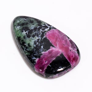 Può includere: Una gemma lucidata con un mix di colori. La pietra ha una base nera con inclusioni verdi e bianche e un prominente motivo rosa a forma di croce. La pietra è liscia e di forma ovale.
