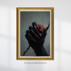 Può includere: Un'opera d'arte incorniciata raffigurante una mano oscura e artigliata che tiene una rosa rosa. Il dipinto ha una cornice dorata ornata e uno sfondo grigio tenue. Il testo "STUDIOINKPRINTS" è in basso.
