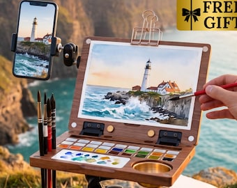 May include: A wooden portable art easel with a built-in watercolor palette, holding a painting of a lighthouse. A smartphone is mounted on the side displaying a reference photo. A hand holds a red paintbrush. Includes a blue tote bag and pencils.