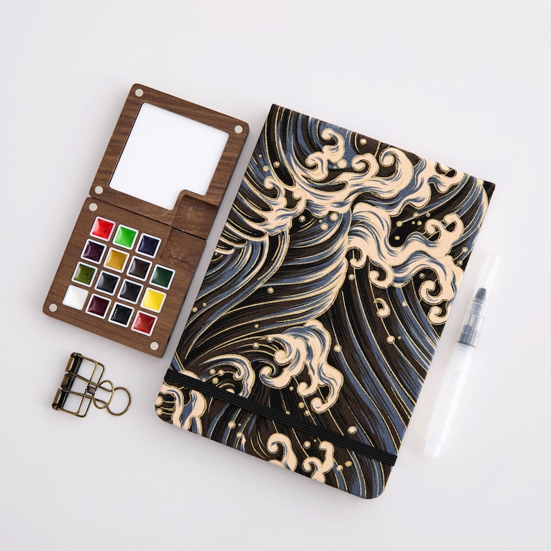 May include: A wooden watercolor palette with twelve paint pans and a mixing area, a notebook featuring a black and beige Japanese-style wave pattern with an elastic closure, and a clear water brush pen with a black cap and a metal binder clip.