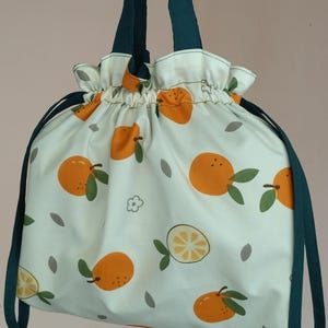 Puede incluir: Una bolsa blanca con cordón con un patrón repetido de cítricos naranjas y hojas verdes. La bolsa tiene asas de color verde oscuro y una parte superior fruncida. El diseño incluye formas de hojas grises y una flor.