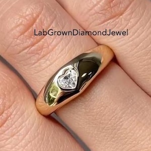 Puede incluir: Un anillo de oro con un diamante en forma de corazón. El anillo tiene una banda lisa y pulida y un engaste de bisel que sujeta el diamante. El texto "LabGrownDiamondJewel" es visible.