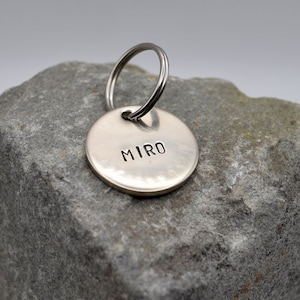 May include: Silver dog tag with a round shape and a keyring. The tag is engraved with the name "MIRO".
